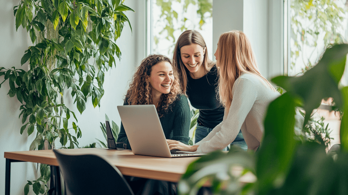 Happy female colleagues collaborating on a project with a laptop in a bright office with plants.