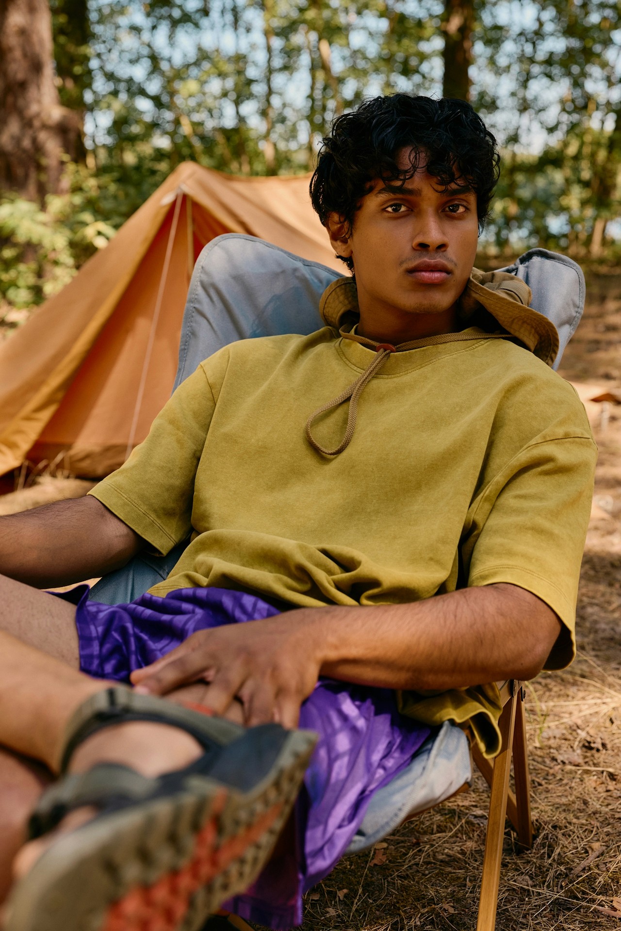 A man seated on a chair in a forest, wearing yellow shirt.