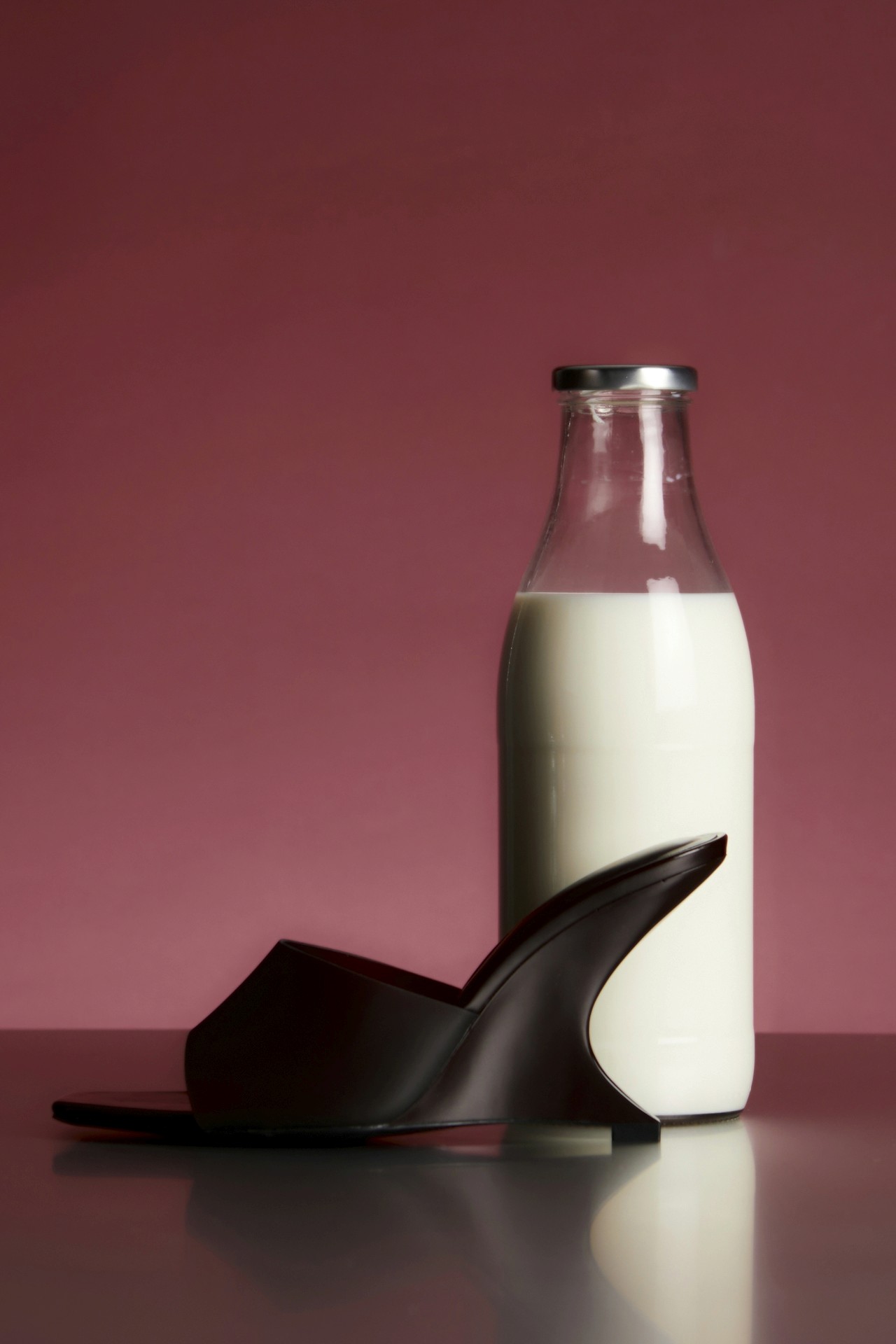 A minimalist still life of a milk bottle and a black high-heeled shoe on a pink background.