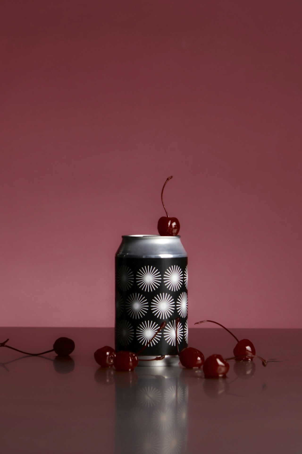 A black can with a cherry placed on top, surrounded by cherries on a reflective surface.