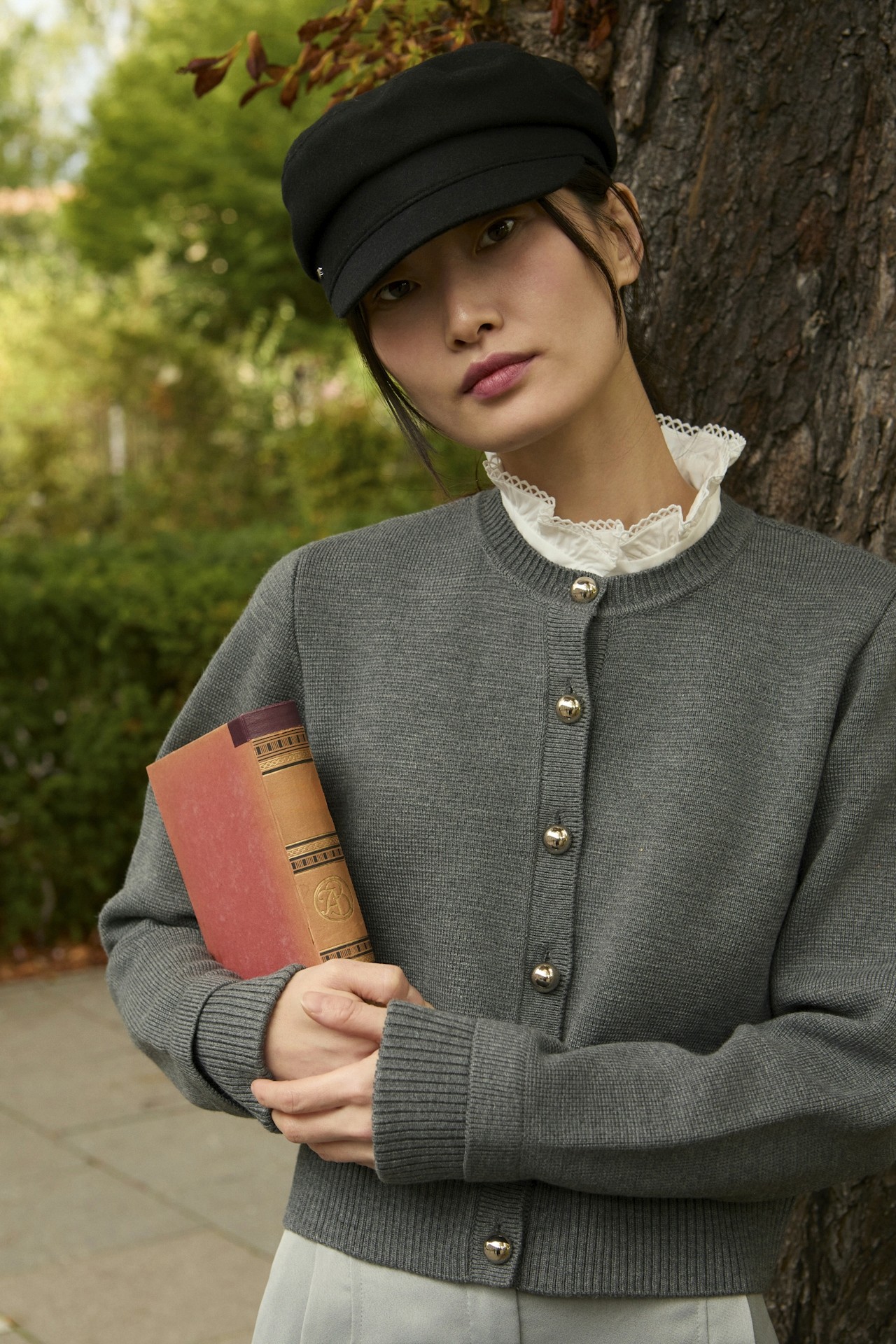 A woman holding a book while standing near a tree in an outdoor setting.