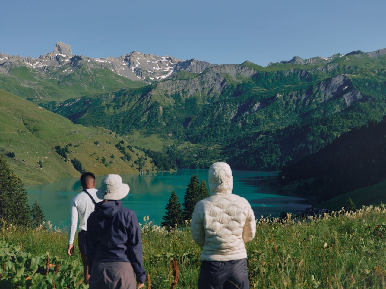 Three friends walking from green mountain slopes toward a lake.