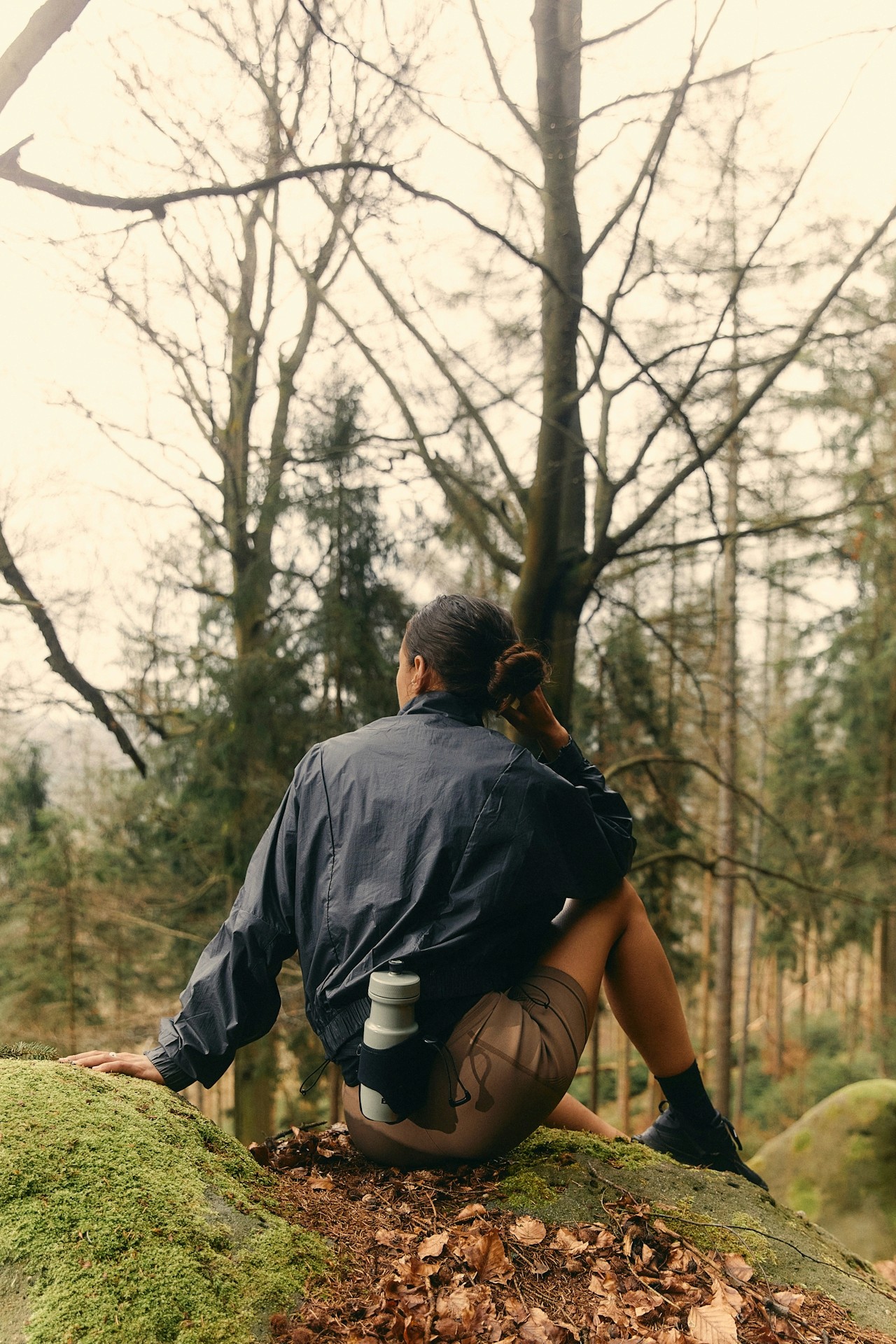 A woman seated on the forest floor among tall trees.