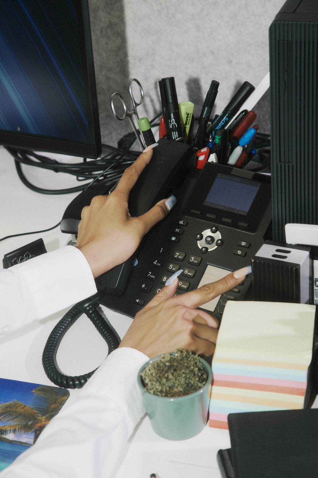 Hands using an office desk phone surrounded by stationery.