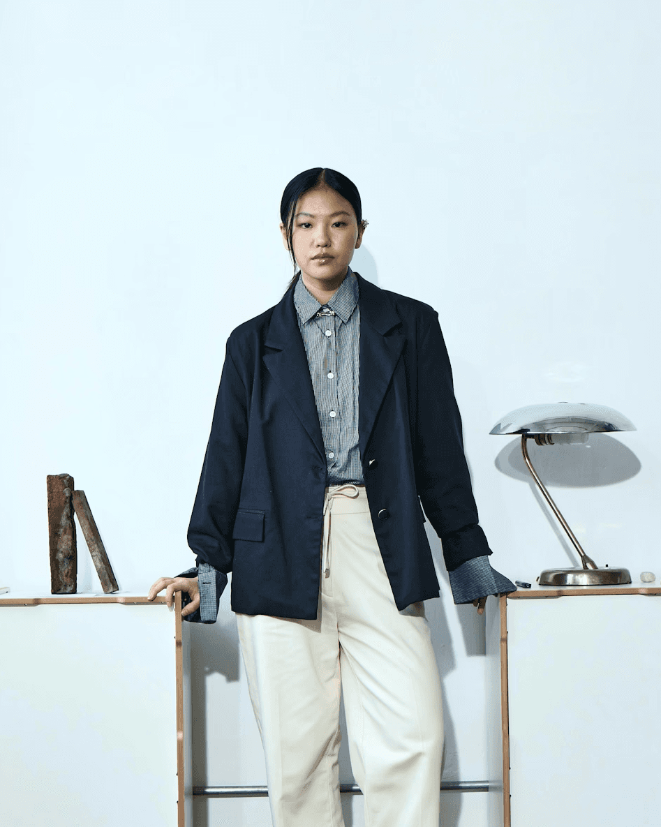 A woman standing confidently in a blazer and trousers in a studio-like workspace.