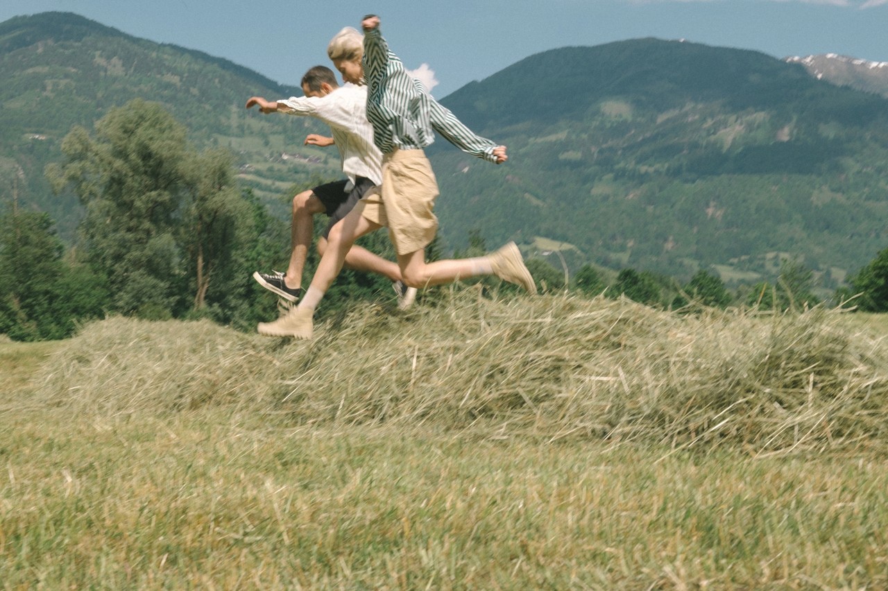 Two people running across a grassy field with mountains behind them.