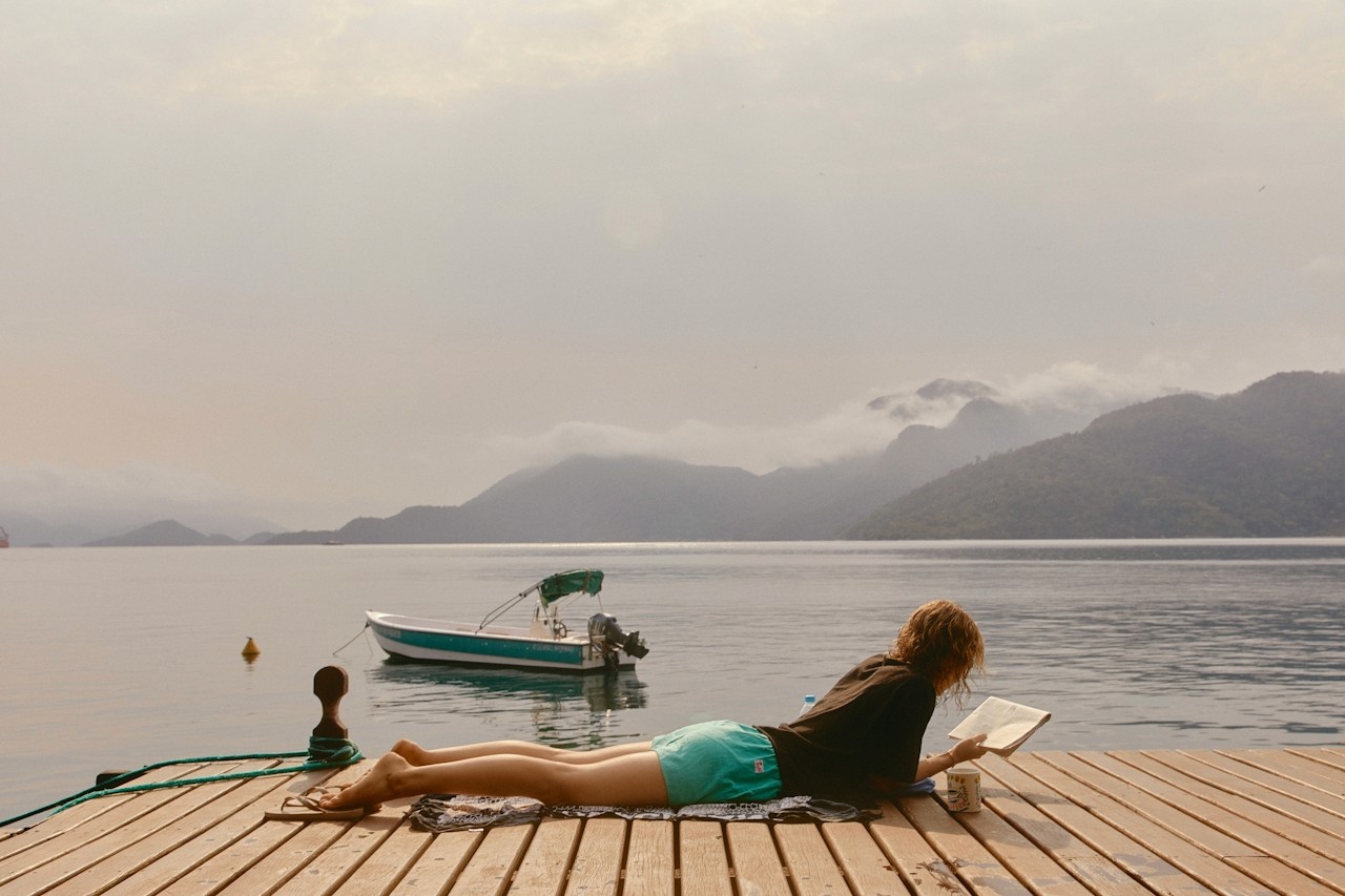 A person relaxing and reading on a dock by calm water and mountains.