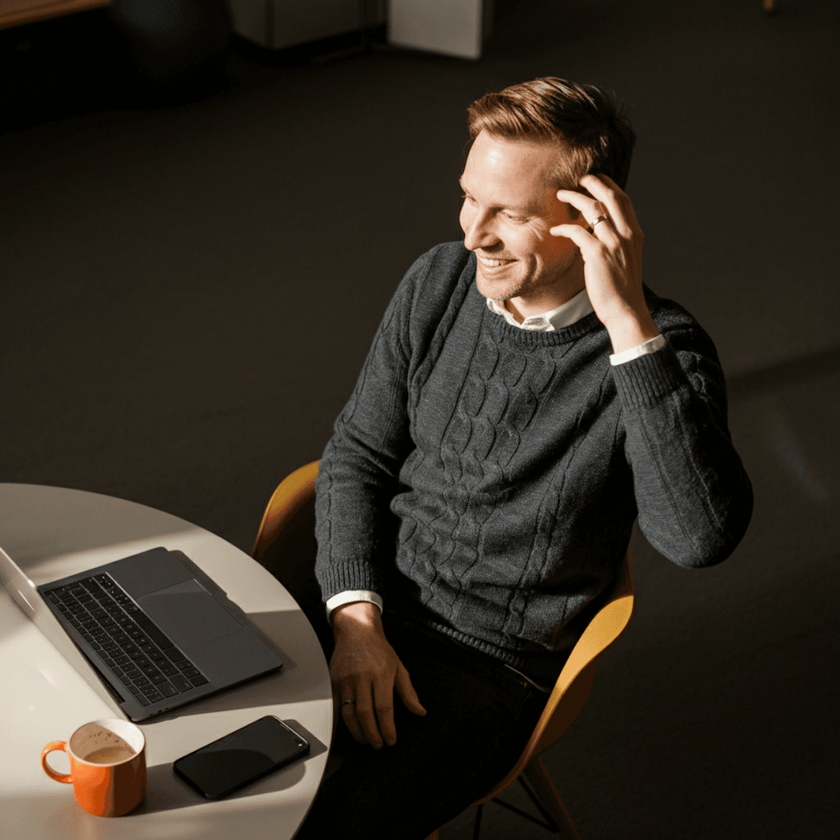 Smiling man sitting at a table with a laptop, phone, and coffee mug, engaged in a video call or online meeting.