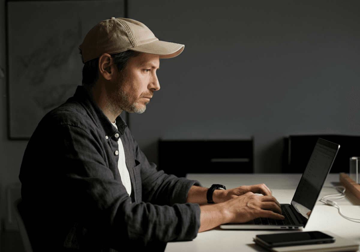 Focused man wearing a cap working on a laptop at a modern desk in a minimal office setting.