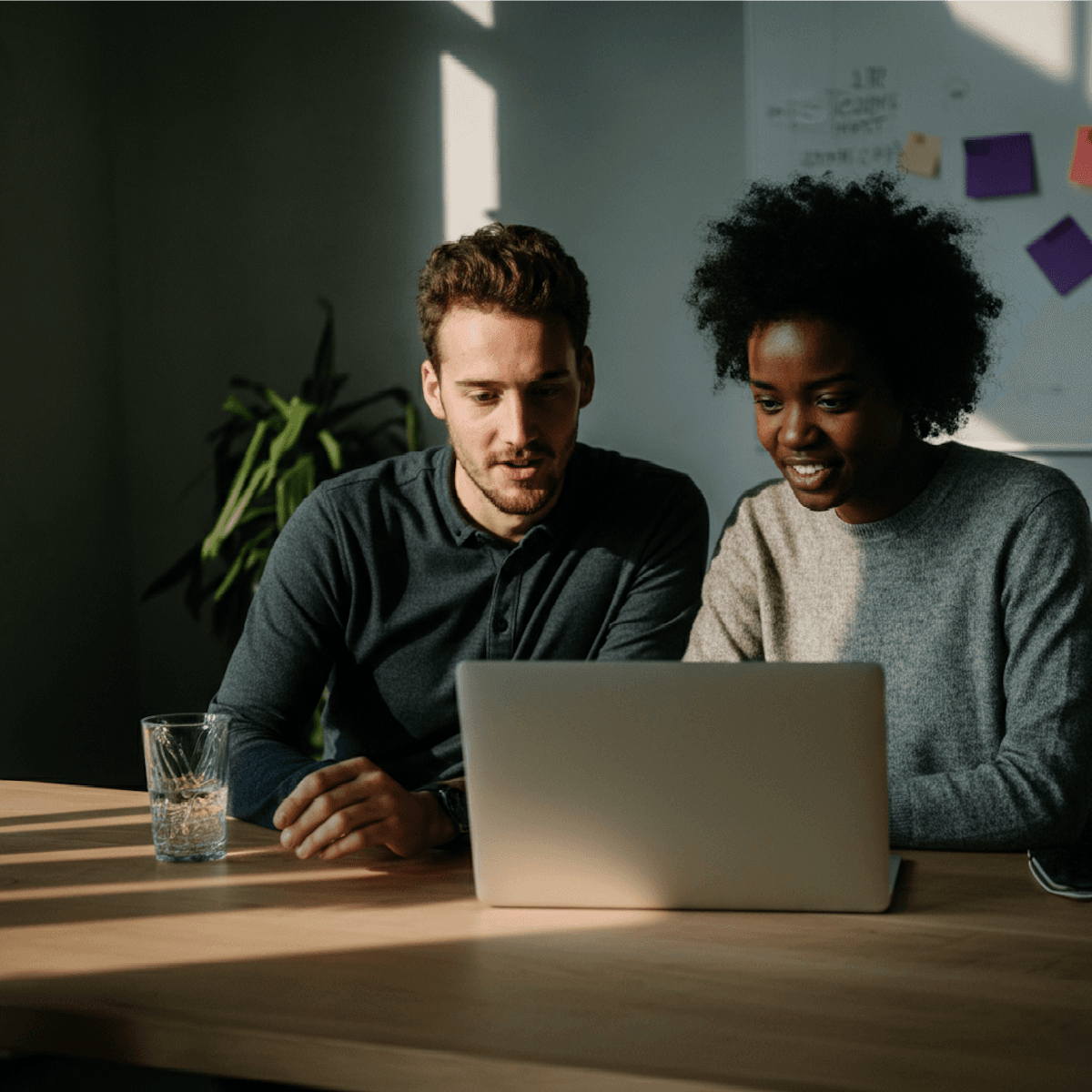 Two colleagues collaborating at a desk, looking at a laptop screen and discussing something with interest.