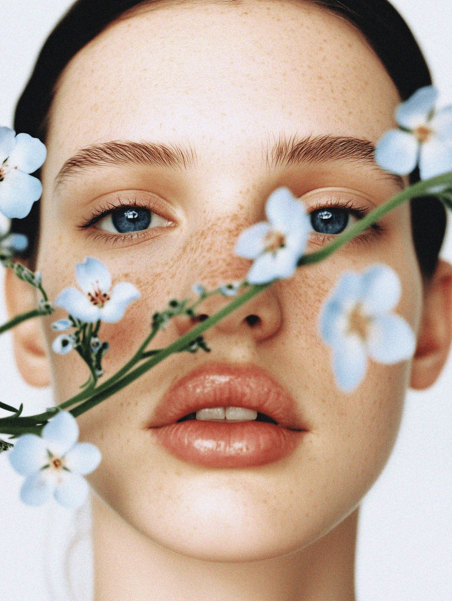 A close-up portrait of a woman with clear, natural skin, delicate freckles, and striking blue eyes, partially obscured by a branch of small, light blue flowers. The image exudes a fresh, natural, and serene aesthetic, perfect for promoting organic skincare, natural beauty products, or floral-inspired cosmetics. This vibrant and soft visual would be beautifully showcased on a **Liqua Premium Ecommerce template**, enhancing the perceived purity and gentle nature of the products.