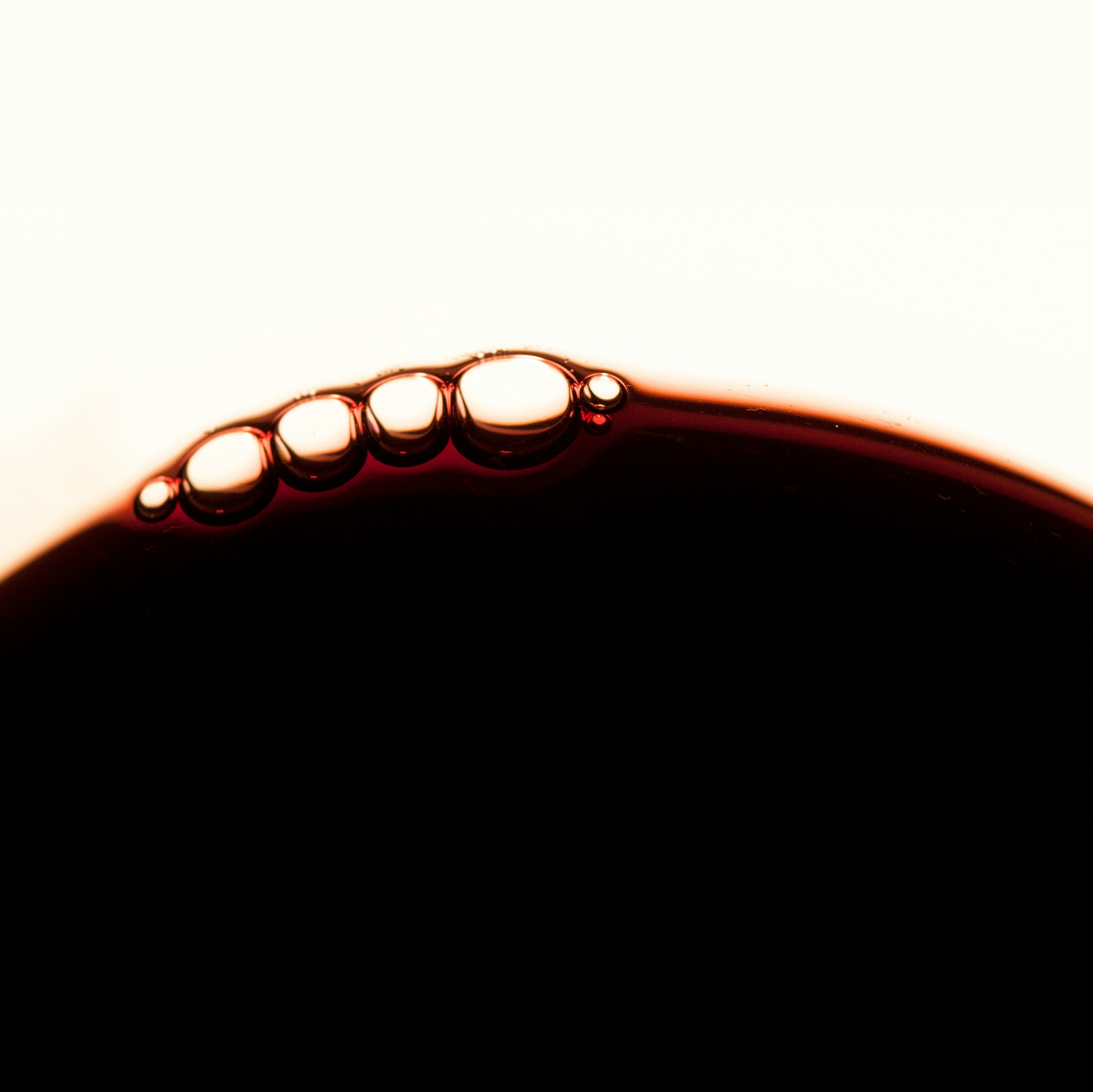 A macro photograph capturing the rich, dark surface of a liquid, likely coffee, wine, or a dark beverage, with a delicate line of five perfectly formed bubbles resting along the top edge. The light background emphasizes the liquid's deep color and the subtle effervescence. This inviting and detailed image is ideal for showcasing gourmet food and beverage products, luxurious bath oils, or high-end cosmetic liquids. It would perfectly enhance product visuals on a **Liqua Premium Ecommerce template**, conveying a sense of quality and sensory appeal.