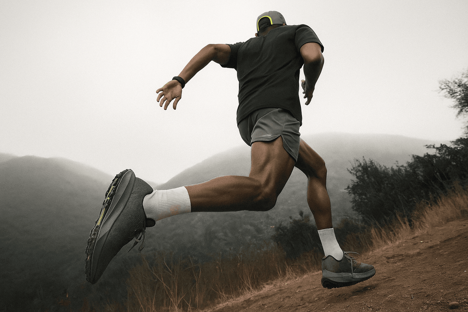 Male athlete in motion leaping forward on a trail path with foggy hills and trees in the background, wearing black shorts and trail shoes.