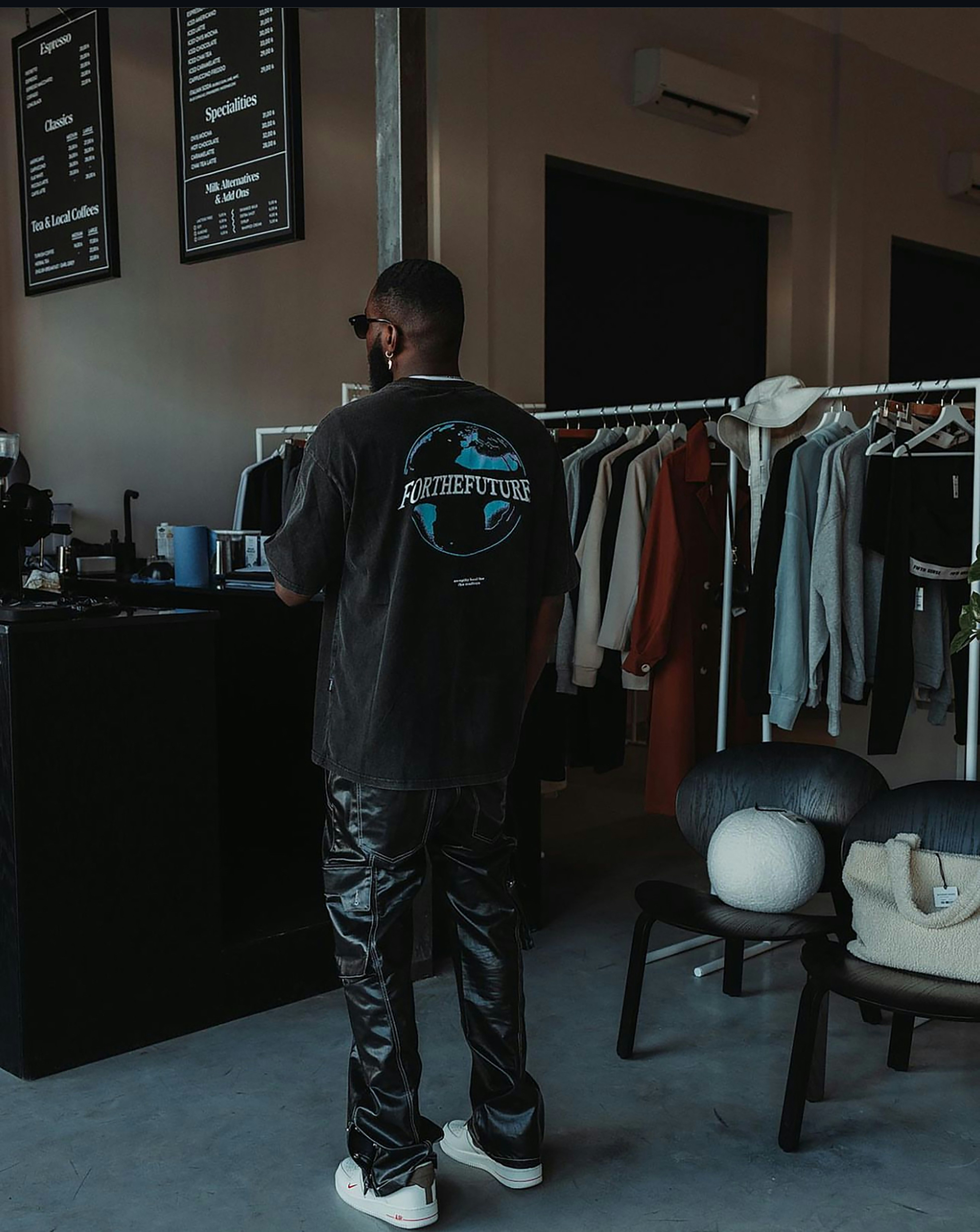 Man from behind in a black "FOR THE FUTURE" globe tee and shiny pants in a stylish cafe and clothing shop.